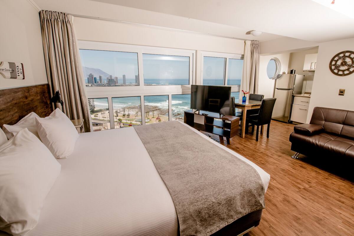 Una habitación de hotel con una cama grande, un escritorio con televisor, un sofá de cuero marrón y vistas a la ciudad y al océano.