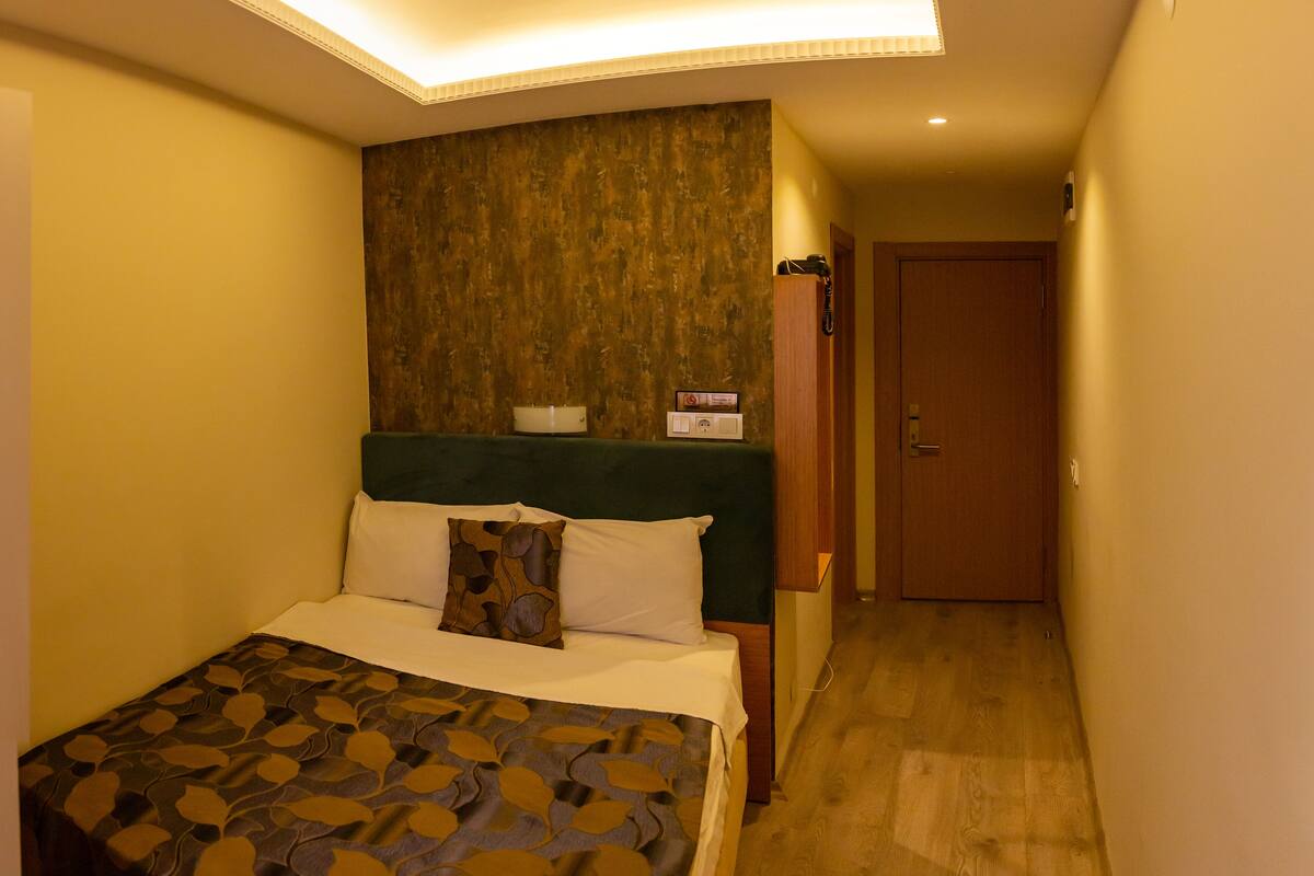 A small, narrow hotel room with a bed, a nightstand, a wall-mounted heater, and a door leading to another room.