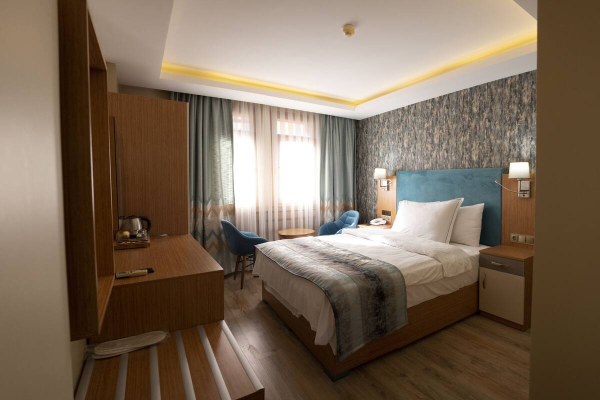 A hotel room with a bed, bedside table, lamp, chair, desk, and curtains.