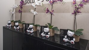 Un área de recepción con un letrero que dice "MGOLDERS GREEN" y varias orquídeas en macetas y ositos de peluche sobre una superficie reflectante.