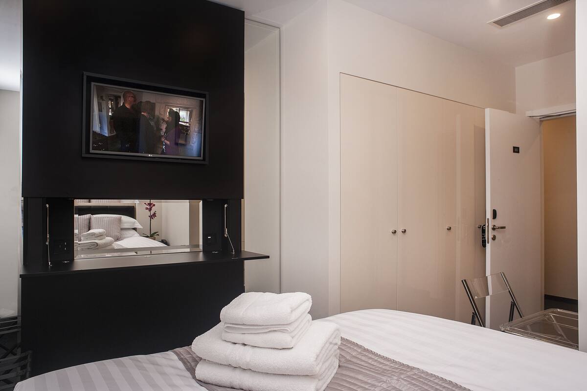 A hotel room with a bed, a black console table with a mirror, a framed picture, and a door with a handle.