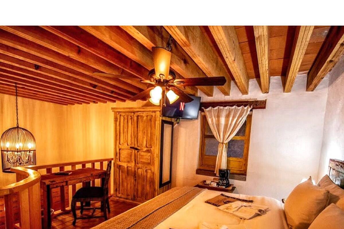 Quarto aconchegante com cama, escrivaninha, cadeira, ventilador de teto e janela com cortinas.