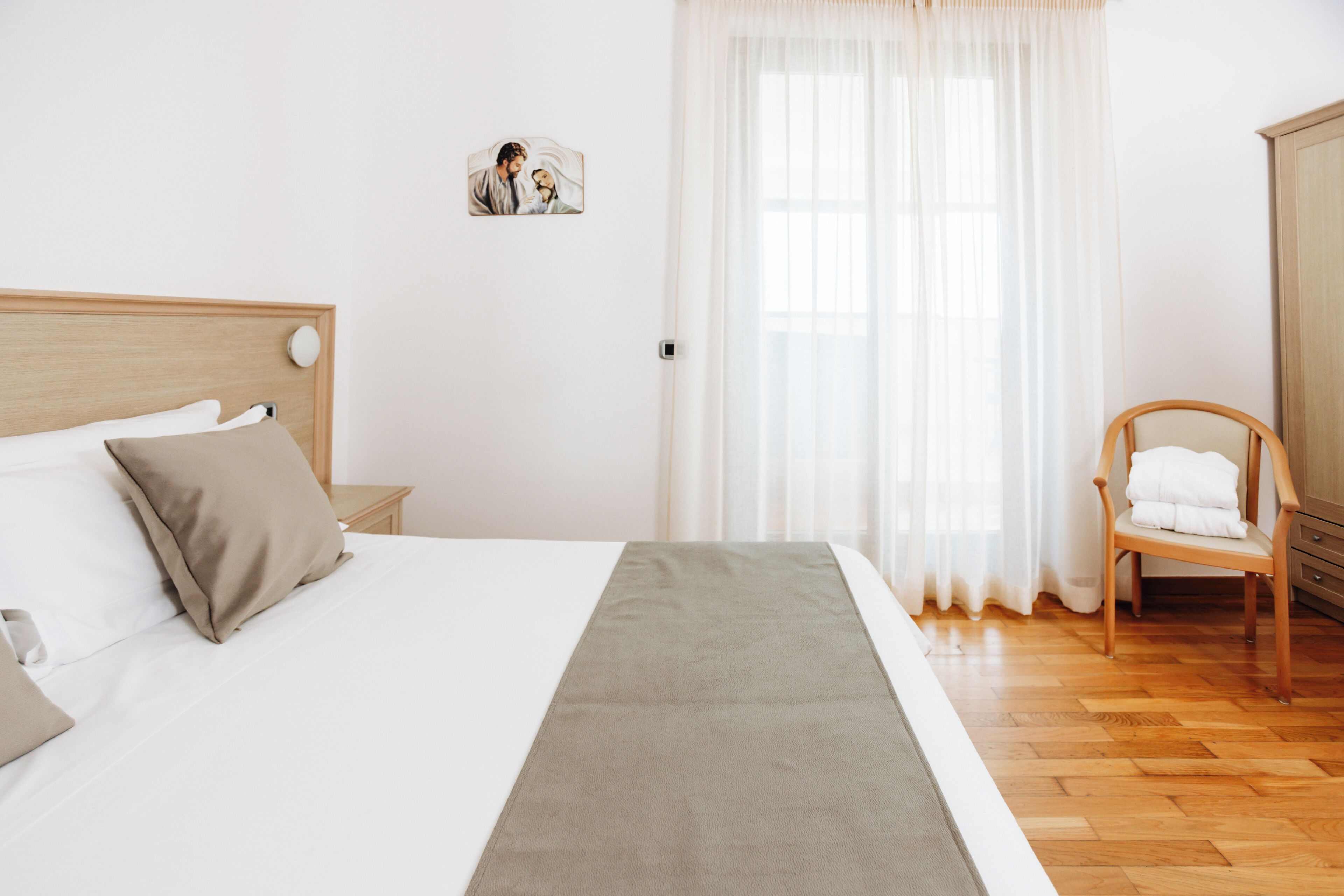 Un letto ben rifatto con una testiera, un comodino, una sedia con un asciugamano e una finestra con tende leggere.