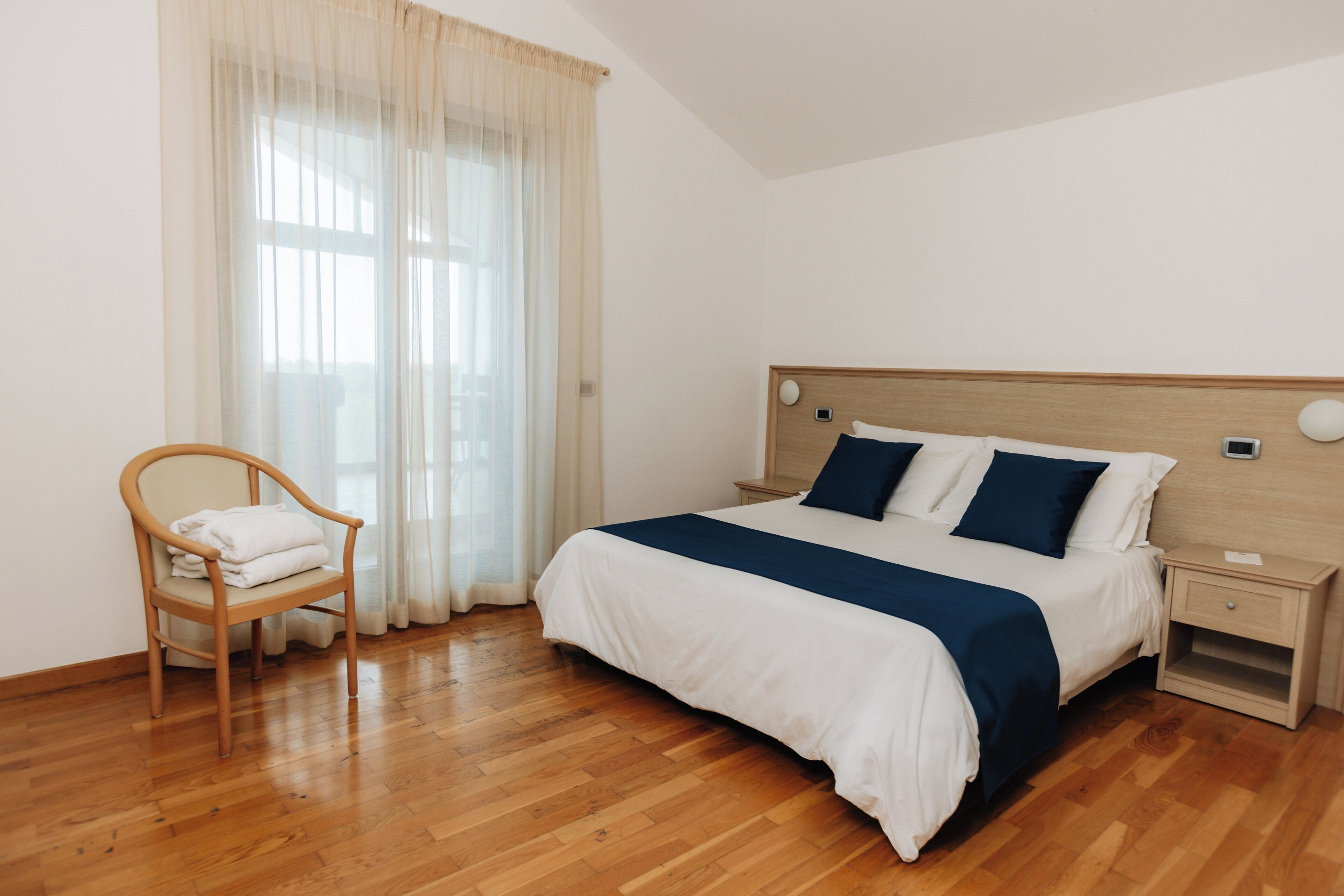Ein Schlafzimmer mit Holzboden, einem Bett mit Holz Kopfteil, einem Nachttisch, einem Stuhl mit Handtuch und einem Fenster mit durchsichtigen Vorhängen.