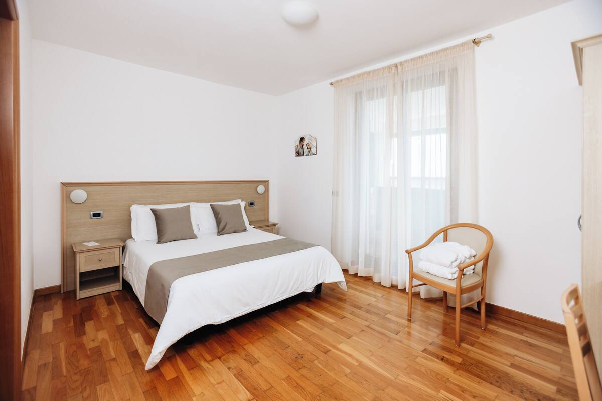 Un dormitorio con piso de madera, una cama con cabecera, una mesita de noche, una silla y una ventana con cortinas translúcidas.