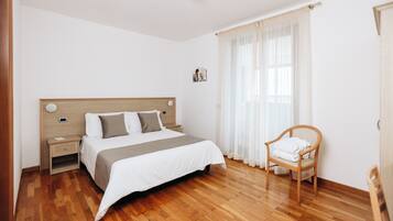 A bedroom with a wooden floor, a bed with a headboard, a nightstand, a chair, and a window with sheer curtains.