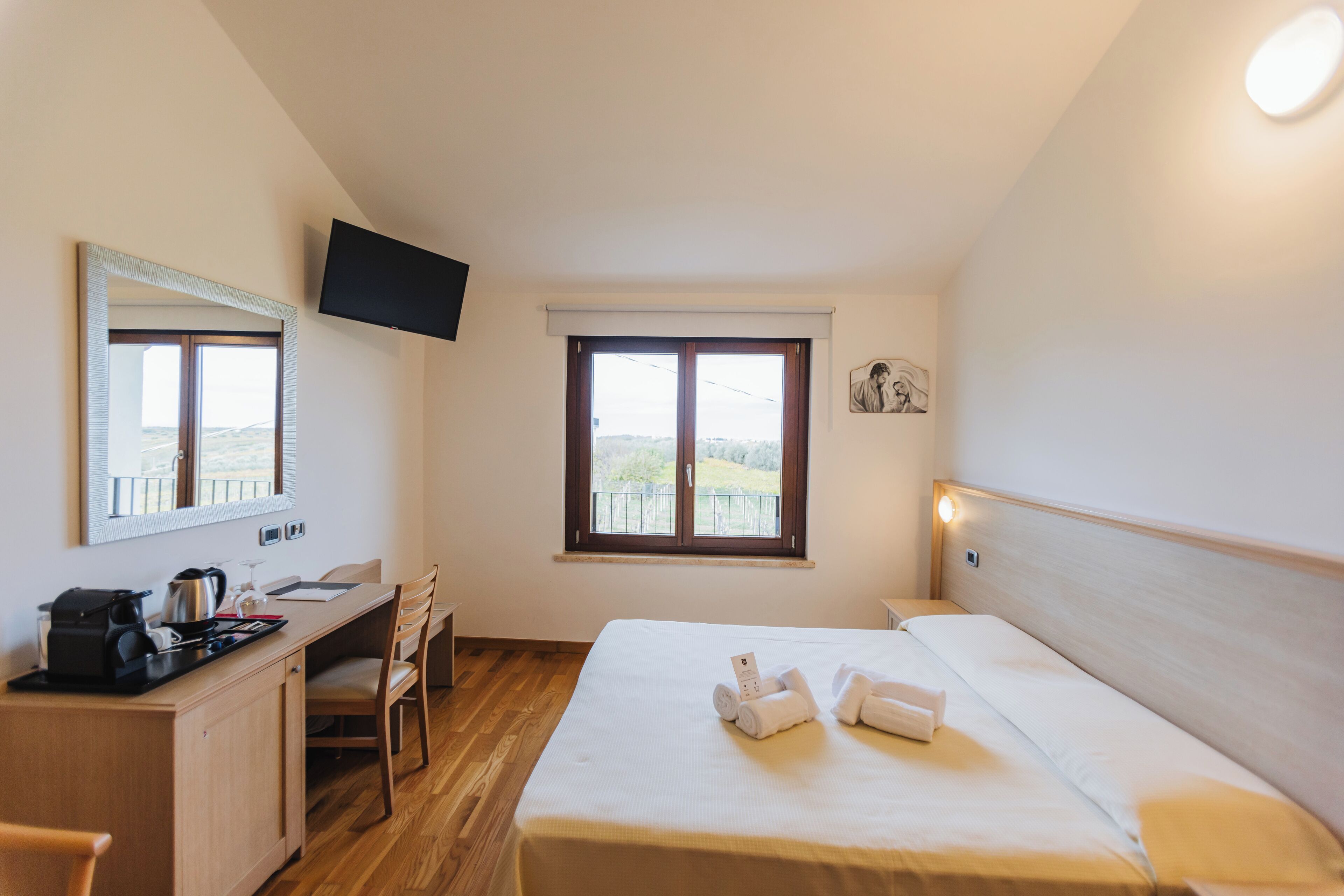 A hotel room with a bed, a desk with a kettle, a chair, a TV mounted on the wall, and a window with a view.