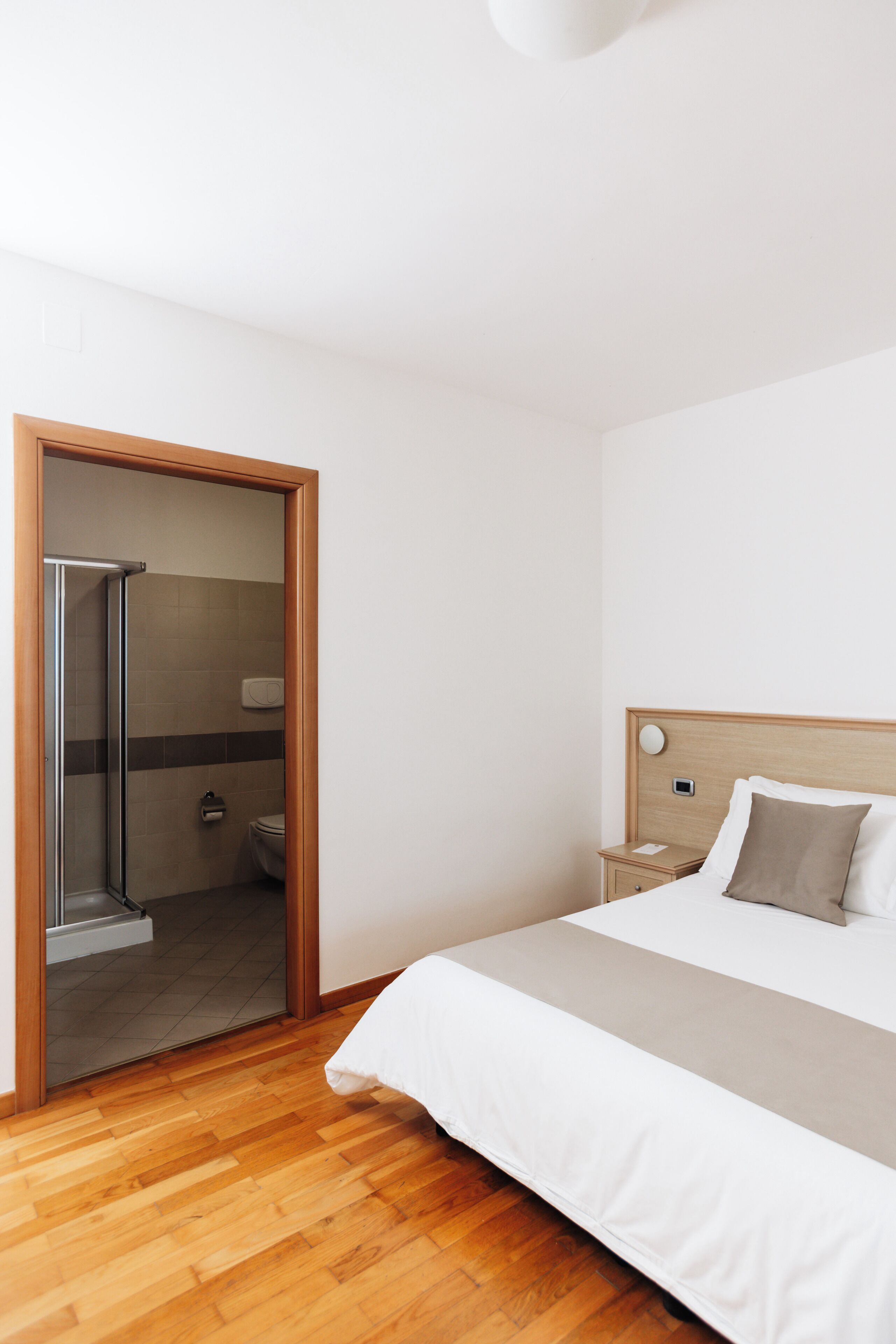 A hotel room with a wooden floor, a bed with white bedding, a wooden headboard, a nightstand, a bedside table, a mirror, and a shower area.