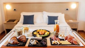 Een ontbijtplateau met croissants, kaas, vleeswaren, fruit en drankjes op een bed in een hotelkamer.