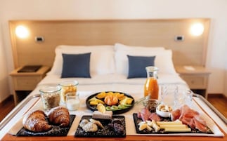 Bandeja de desayuno con croissants, queso, embutidos, frutas y bebidas sobre una cama en una habitación de hotel.
