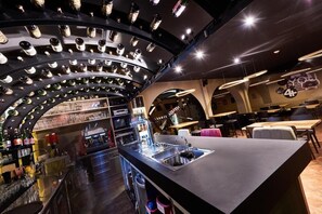 Zona de bar moderna con una estantería curva que exhibe botellas, una barra con lavamanos y áreas de descanso con mesas y sillas.