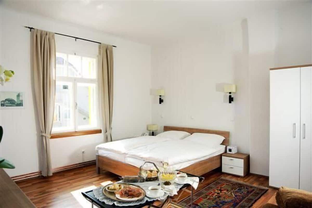 Een hotelkamer met een bed, nachtkastjes, een kledingkast, een raam met gordijnen en een ontbijttafel op de vloer.
