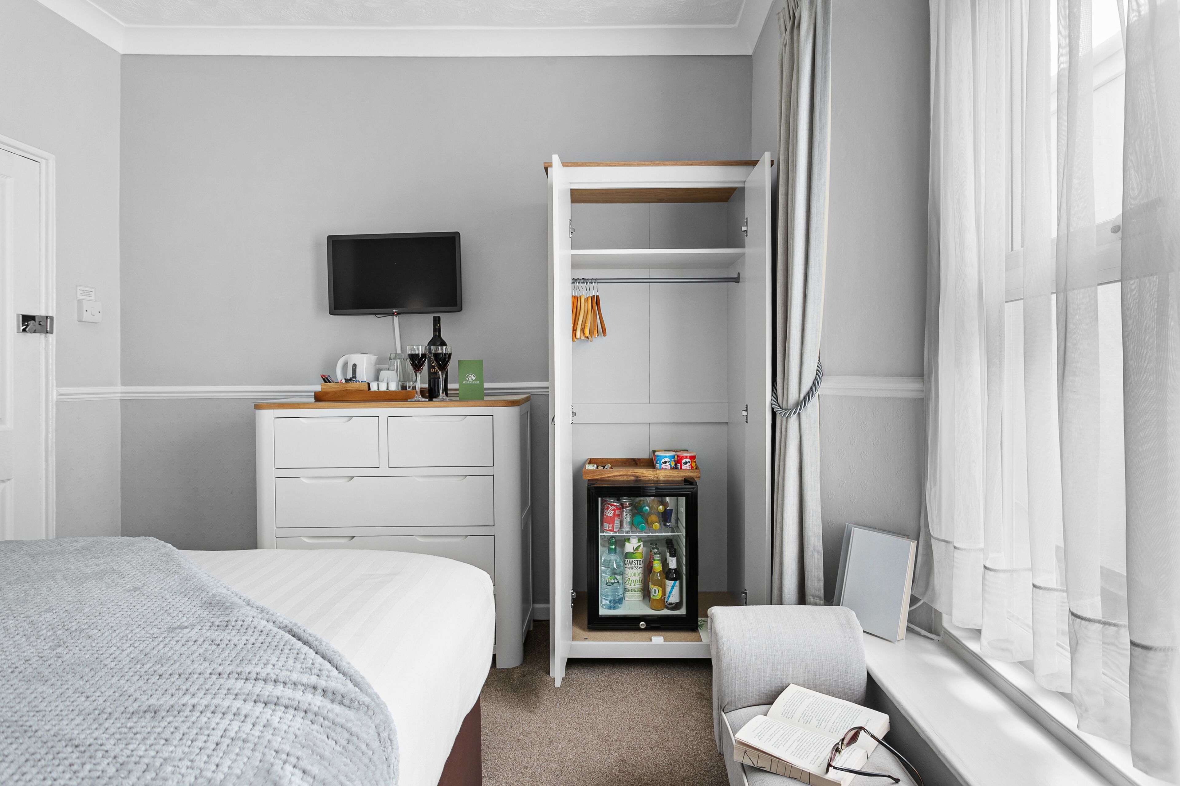 A hotel room with a bed, a bedside table with a lamp, a television, a minibar, and a window with curtains.