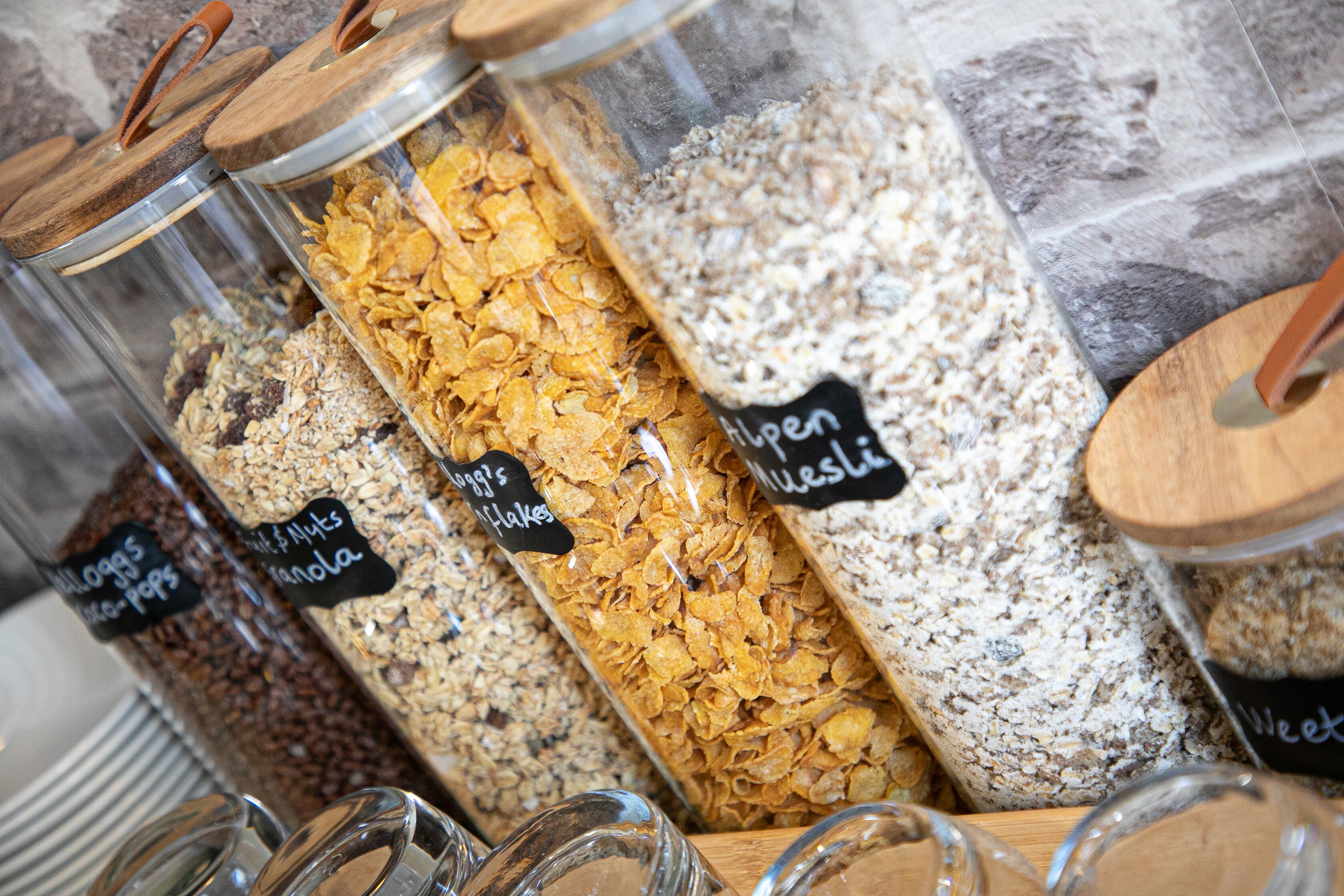 Variedad de barras de granola y cereales en recipientes transparentes con tapas de madera, etiquetadas con nombres como 