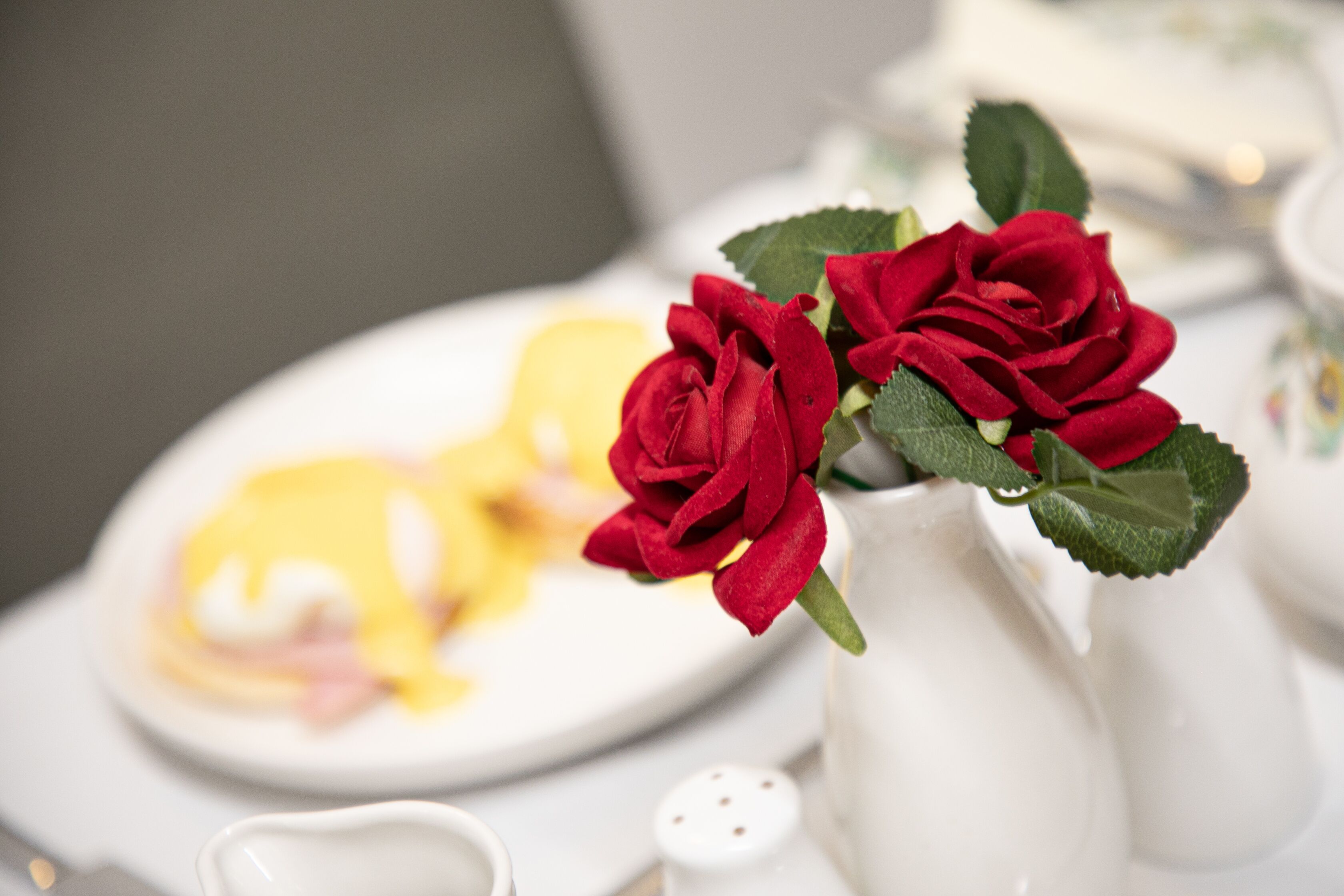 A table with a white vase containing red roses, a white plate with yellow food, and a salt shaker.