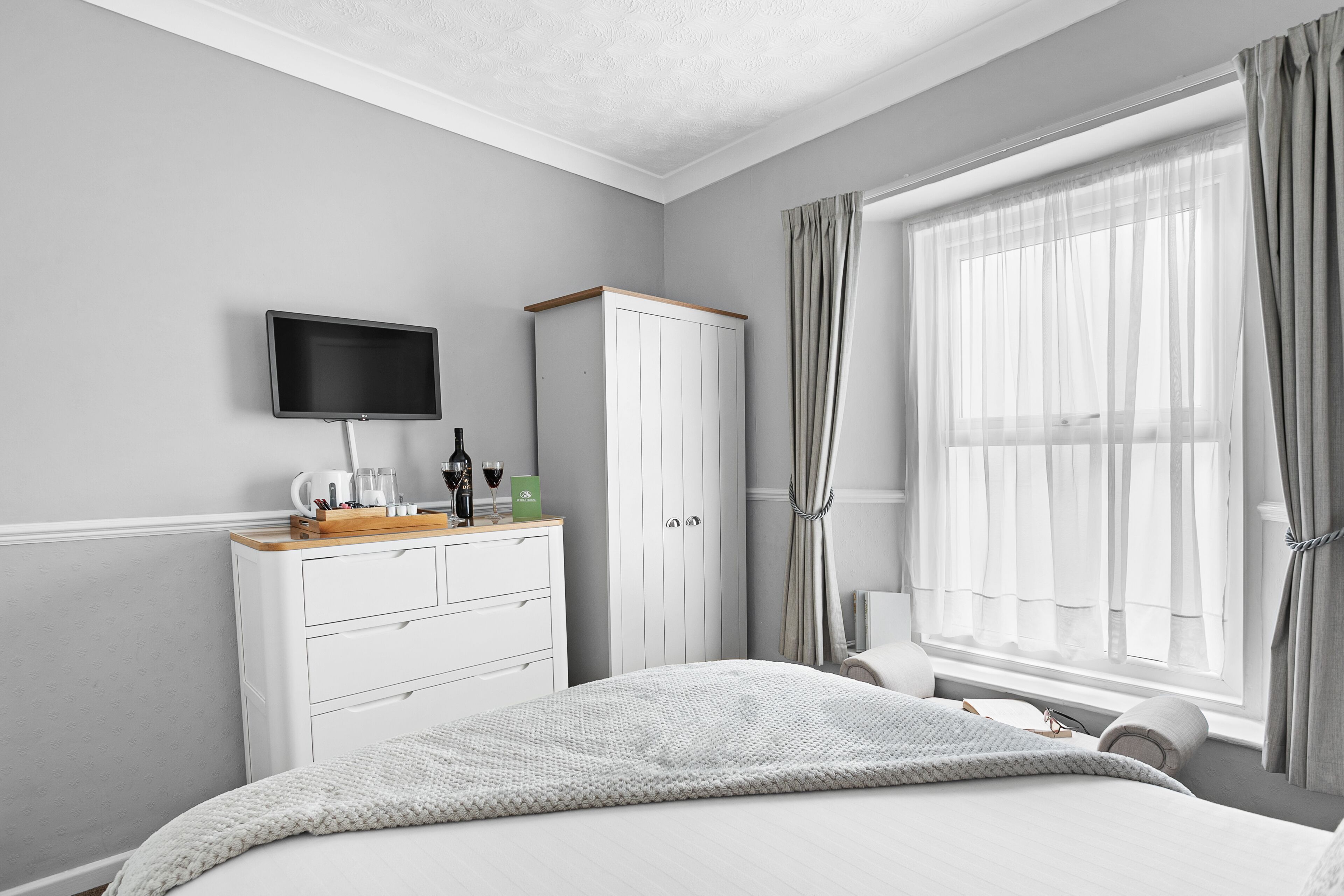 A bedroom with a bed, a TV mounted on the wall, a wardrobe, and a window with curtains.