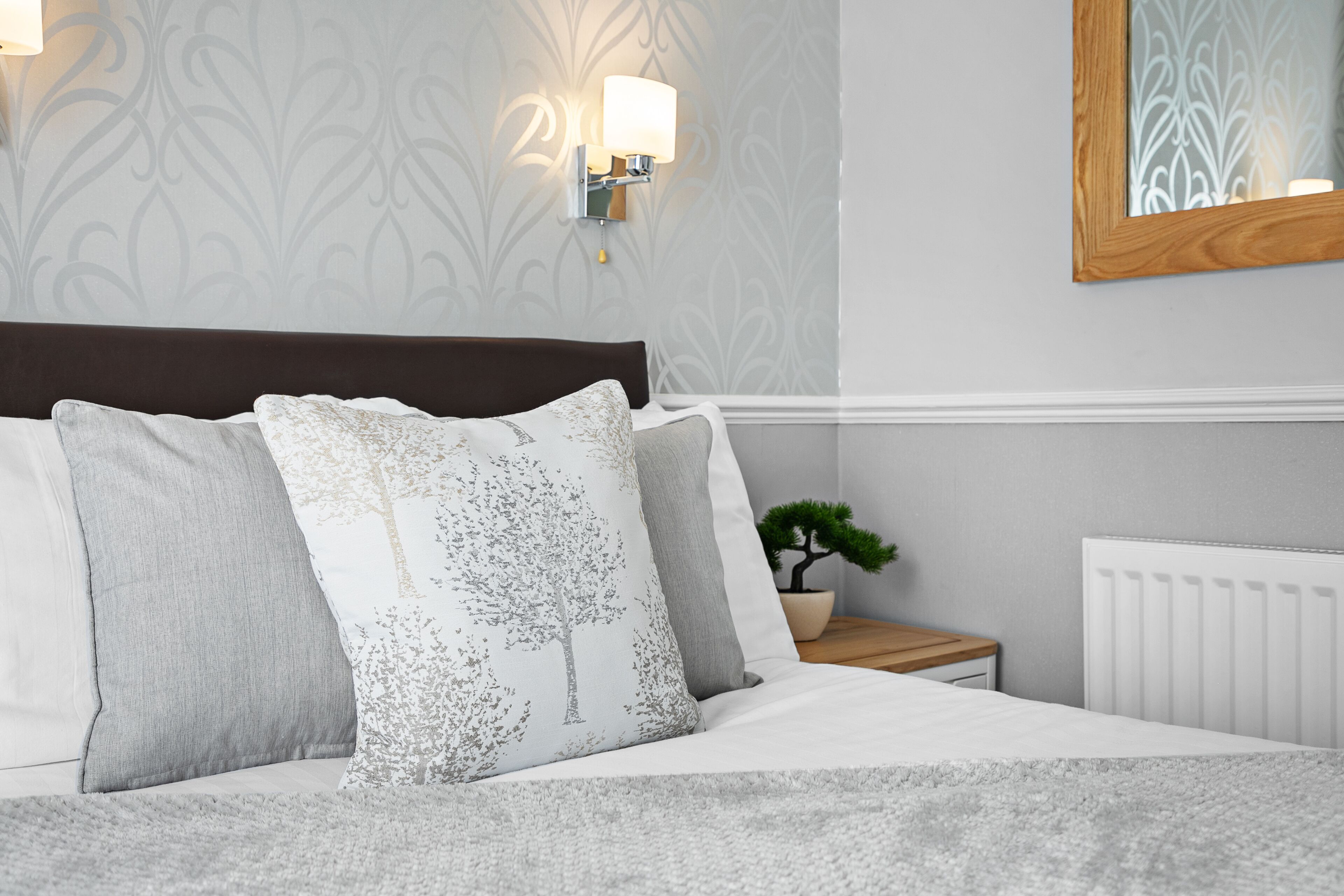 A neatly made bed with a white and gray patterned pillow, a small potted plant, and a wall-mounted light fixture.