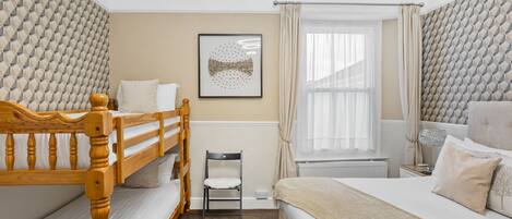 A bedroom with a bunk bed, a single bed, a chair, a window with curtains, and a framed picture on the wall.