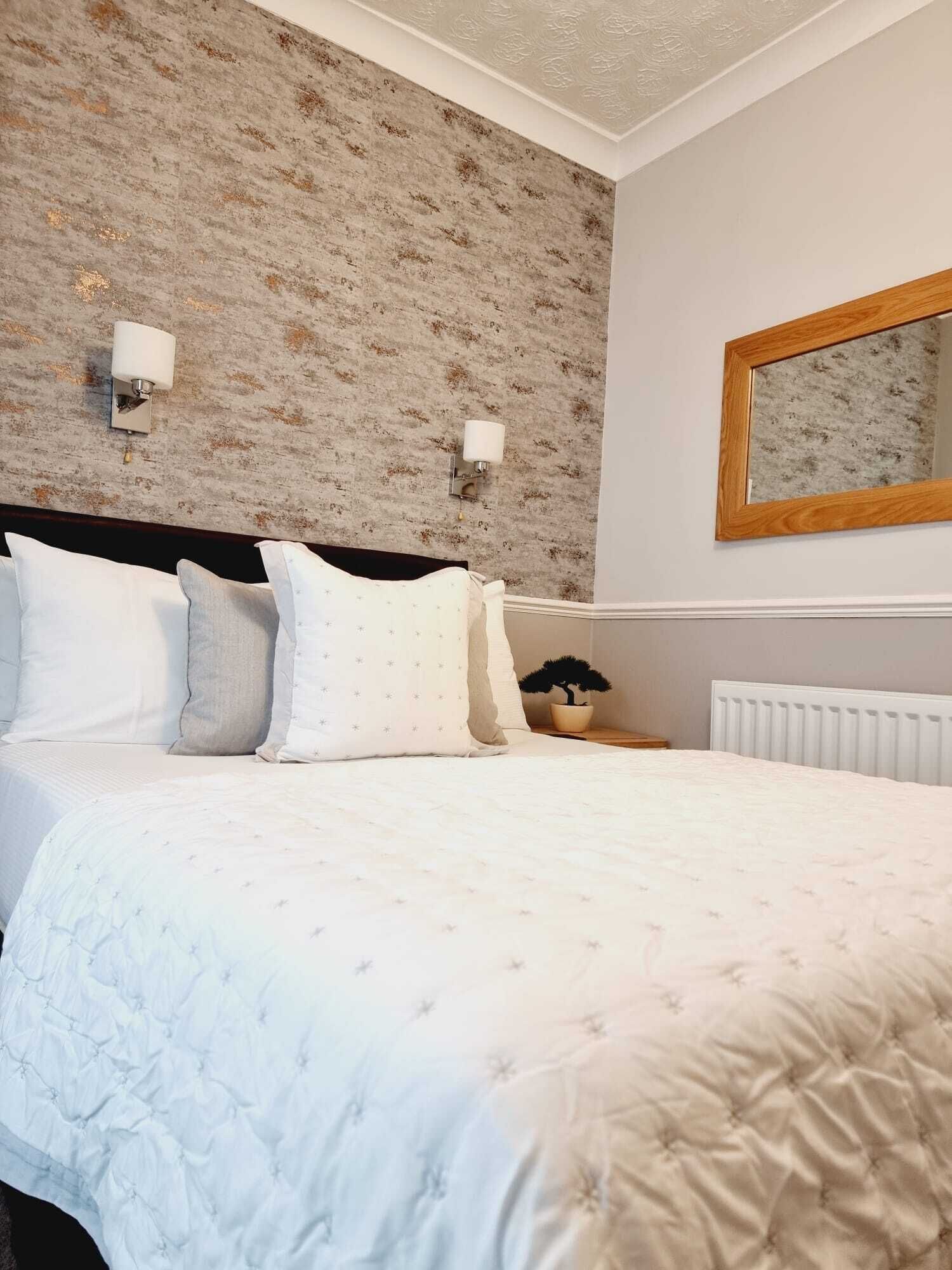 A neatly made bed with white bedding and multiple pillows, a wooden headboard, a wall-mounted mirror, and a radiator.