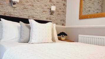 A neatly made bed with white bedding and multiple pillows, a wooden headboard, a wall-mounted mirror, and a radiator.