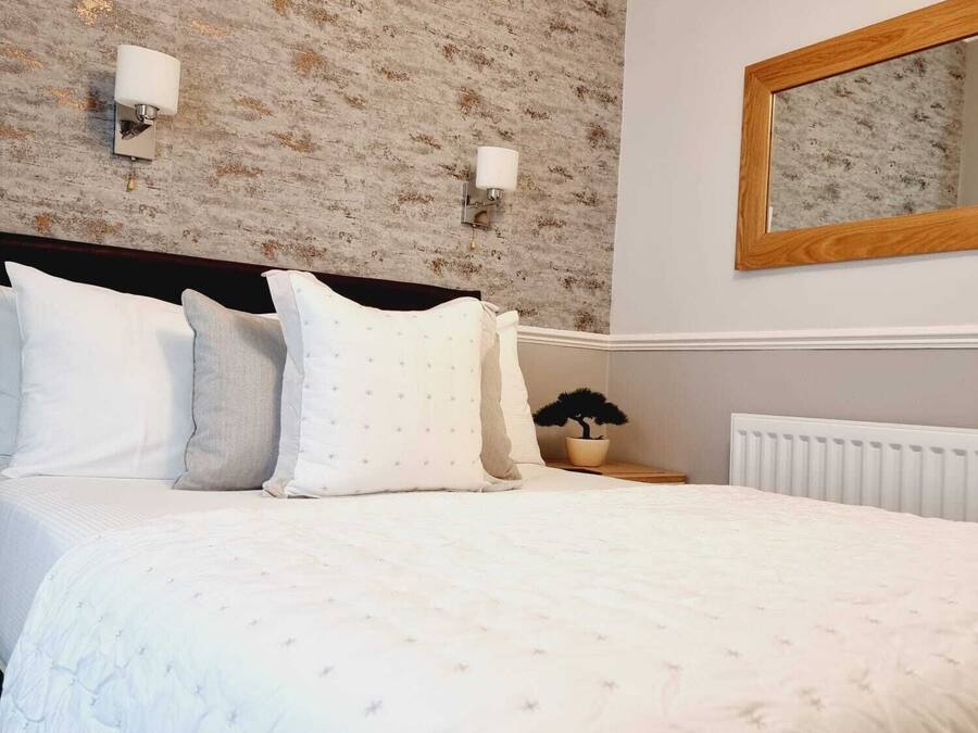 Een net opgemaakt bed met witte beddengoed en meerdere kussens, een houten hoofdbord, een wandspiegel en een radiator.