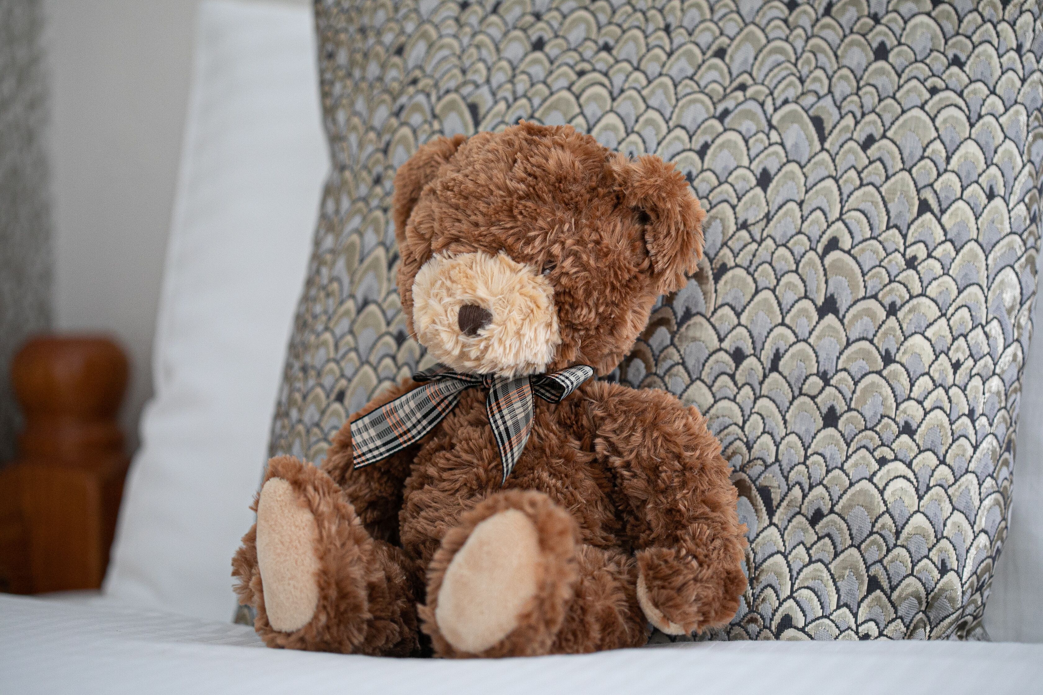 Un orsetto di peluche seduto su un letto con un cuscino decorato sullo sfondo.