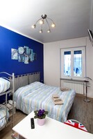 Un dormitorio con una cama individual alta, una mesita de noche con un control remoto y una planta en maceta, una ventana con cortinas y una lámpara en el techo.
