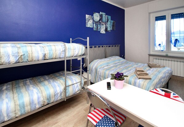 Habitación tipo dormitorio con literas, escritorio, silla, planta en maceta, control remoto y ventana con cortinas.