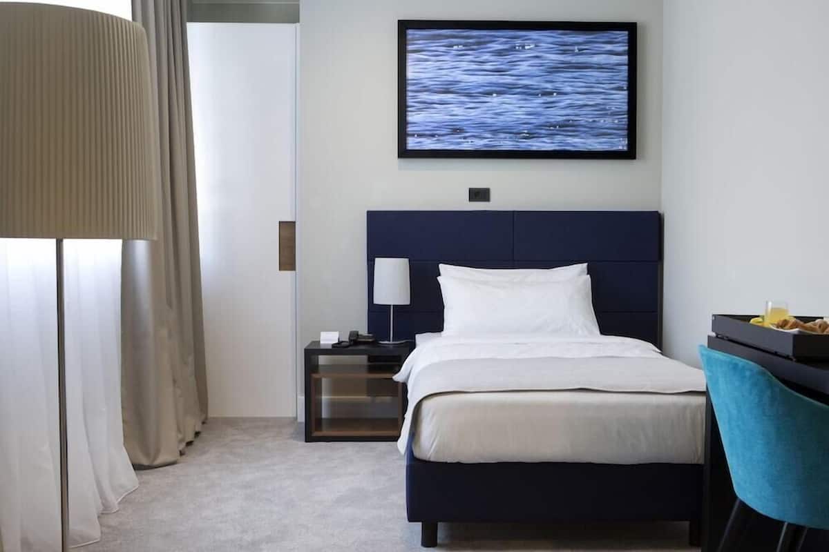 Una habitación de hotel con una cama, una mesita de noche, un televisor montado en la pared, un escritorio con una silla y una ventana con cortinas.