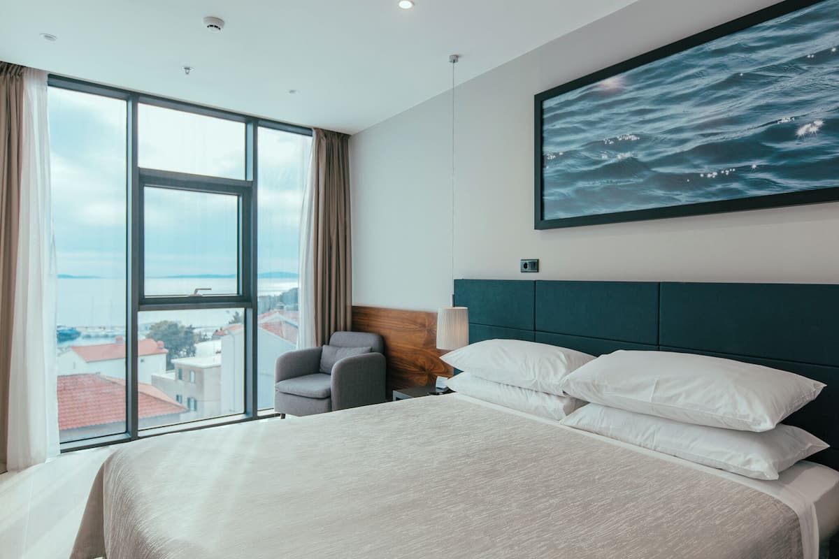 Habitación de hotel con una cama grande, vistas de la ciudad a través de una ventana, una silla y un cuadro con olas en la pared.