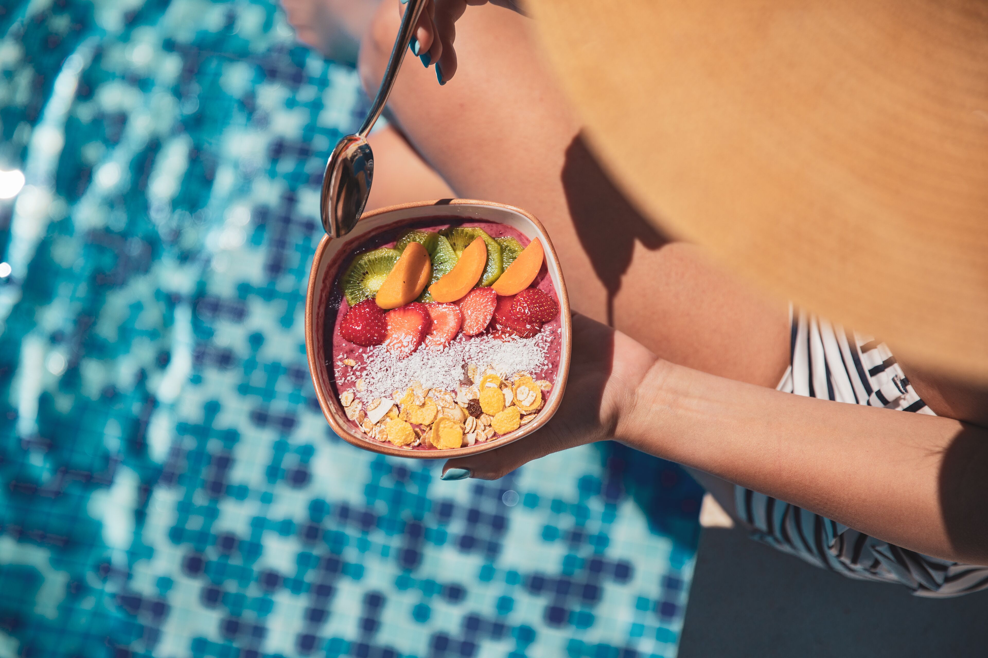 A person holding a bowl of fruit salad with strawberries, kiwi, and other fruits, near a pool.