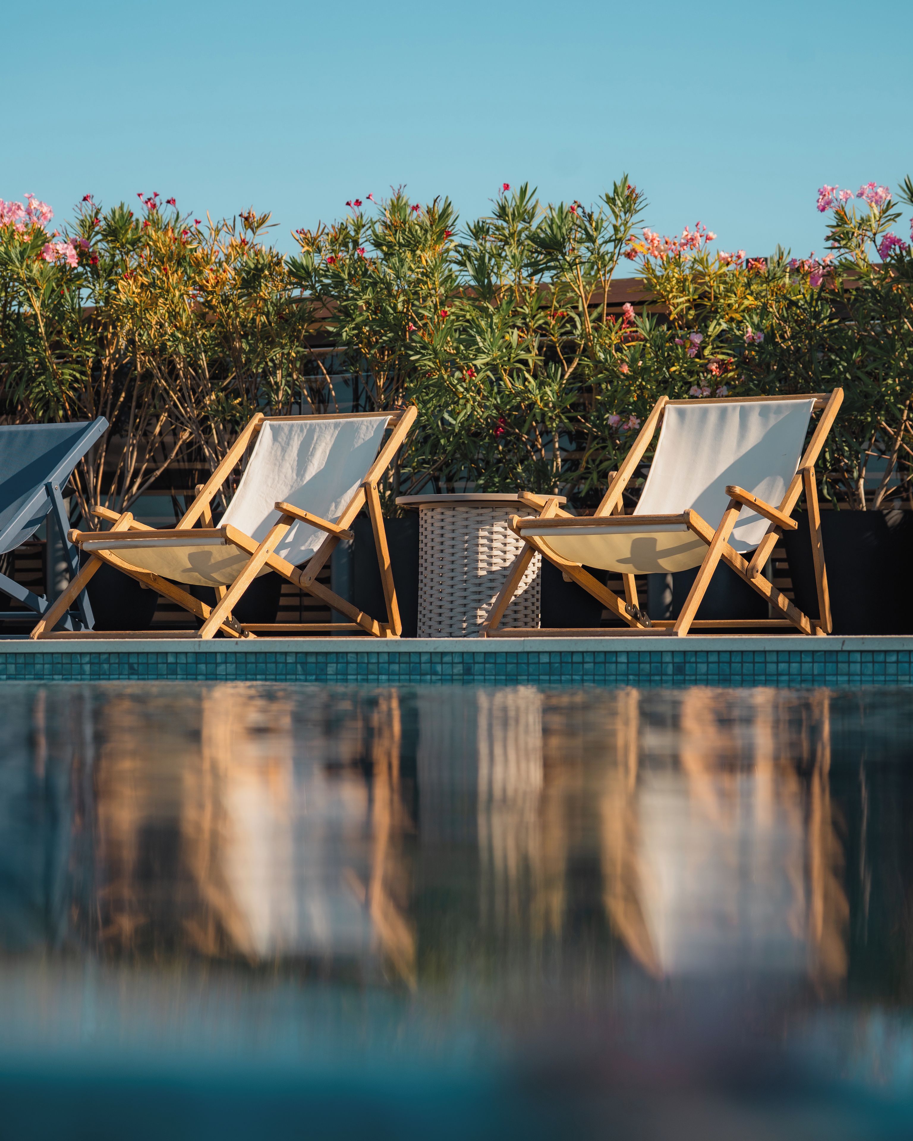 Two wooden lounge chairs by a poolside with a tiled edge and a backdrop of flowering shrubs.