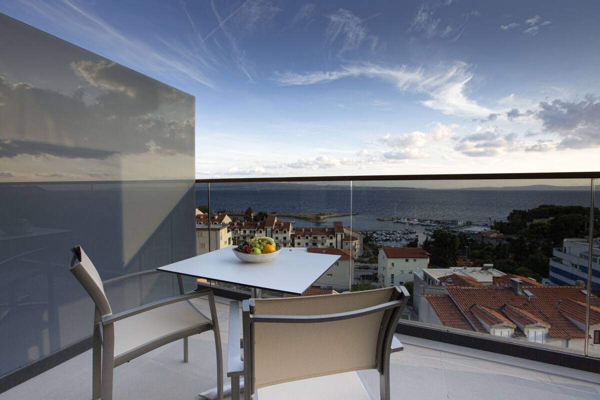 Un balcón con mesa y sillas, con vistas a una ciudad costera que incluye edificios y un puerto deportivo.