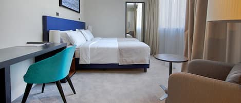 A hotel room with a bed, a desk, a chair, a small table, and a TV mounted on the wall.