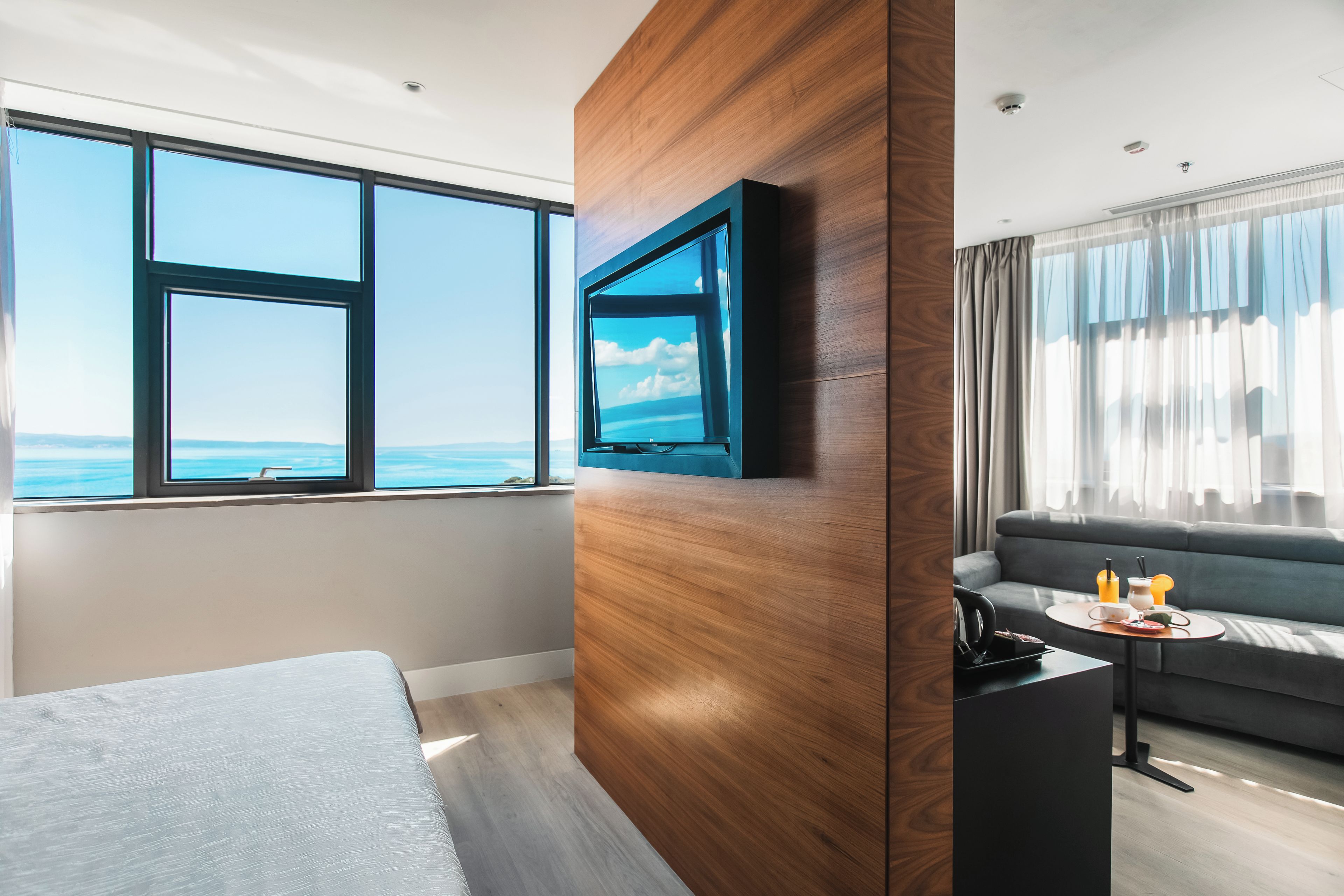 A modern hotel room with a large window, a bed, a TV, a sofa, and a small table with a tray.