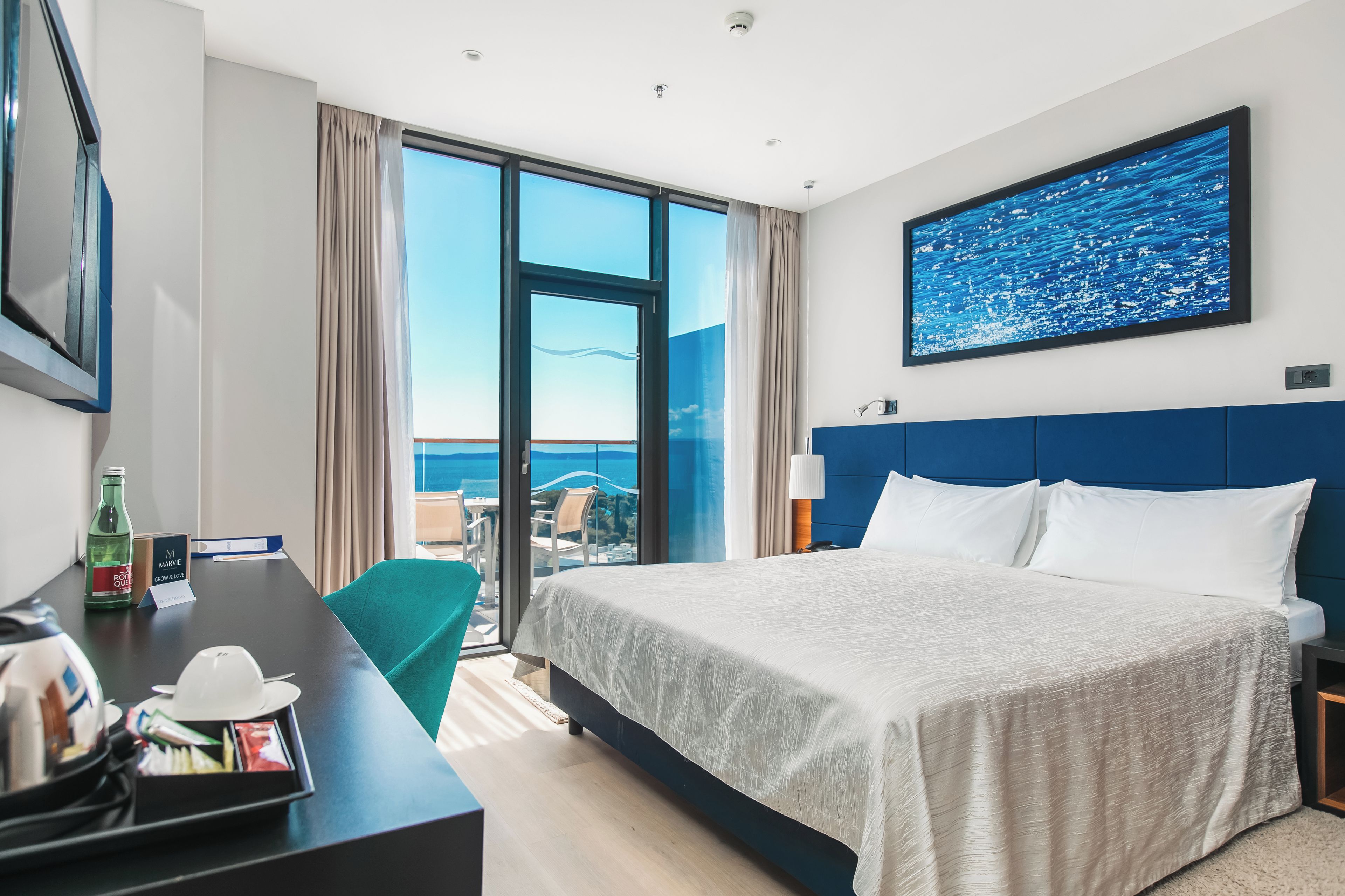 Une chambre d’hôtel avec un grand lit, un bureau avec une chaise, une vue sur la mer et un tableau au mur.