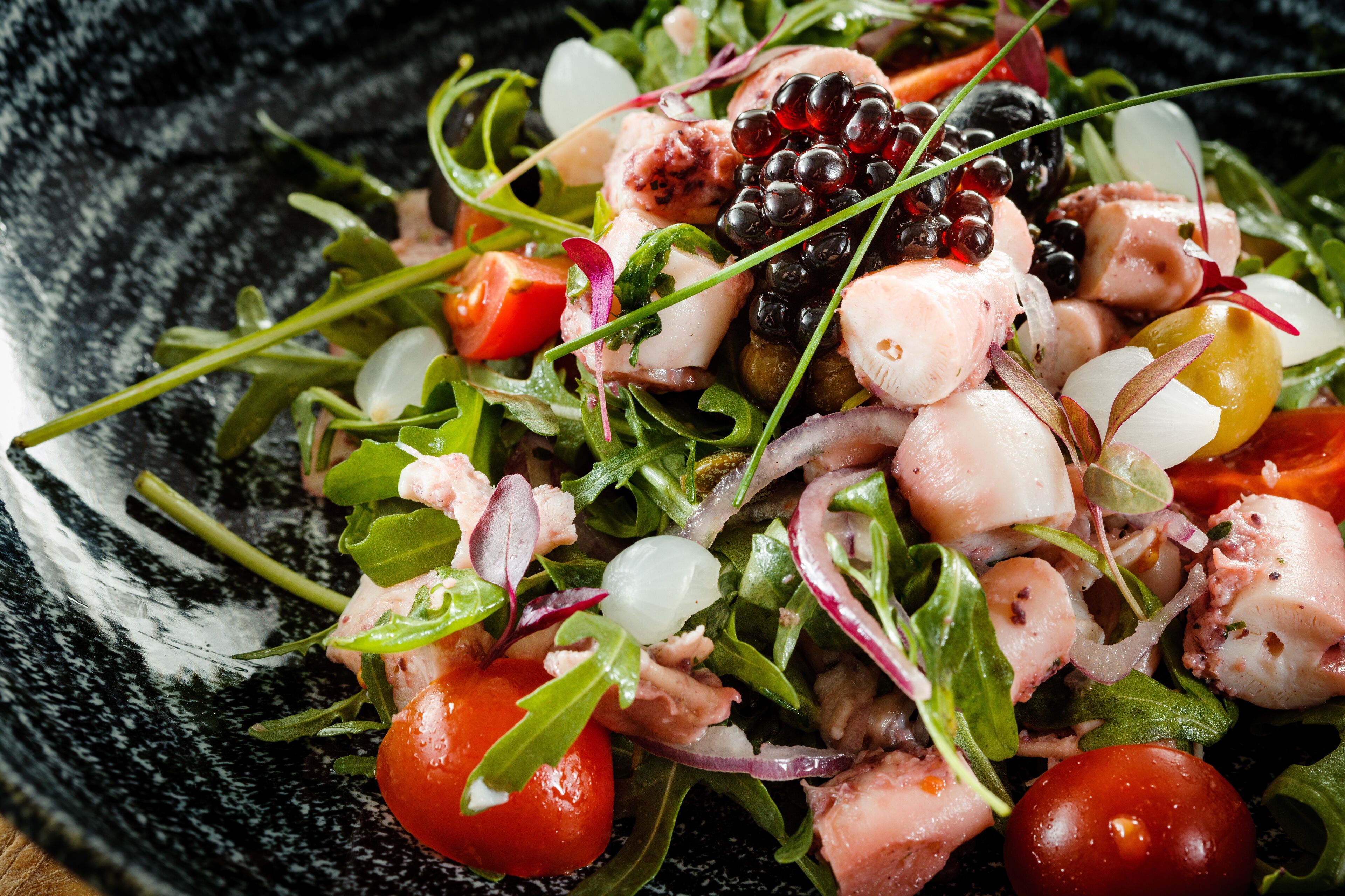 A salad with various ingredients including tomatoes, greens, and possibly seafood, served on a black plate.