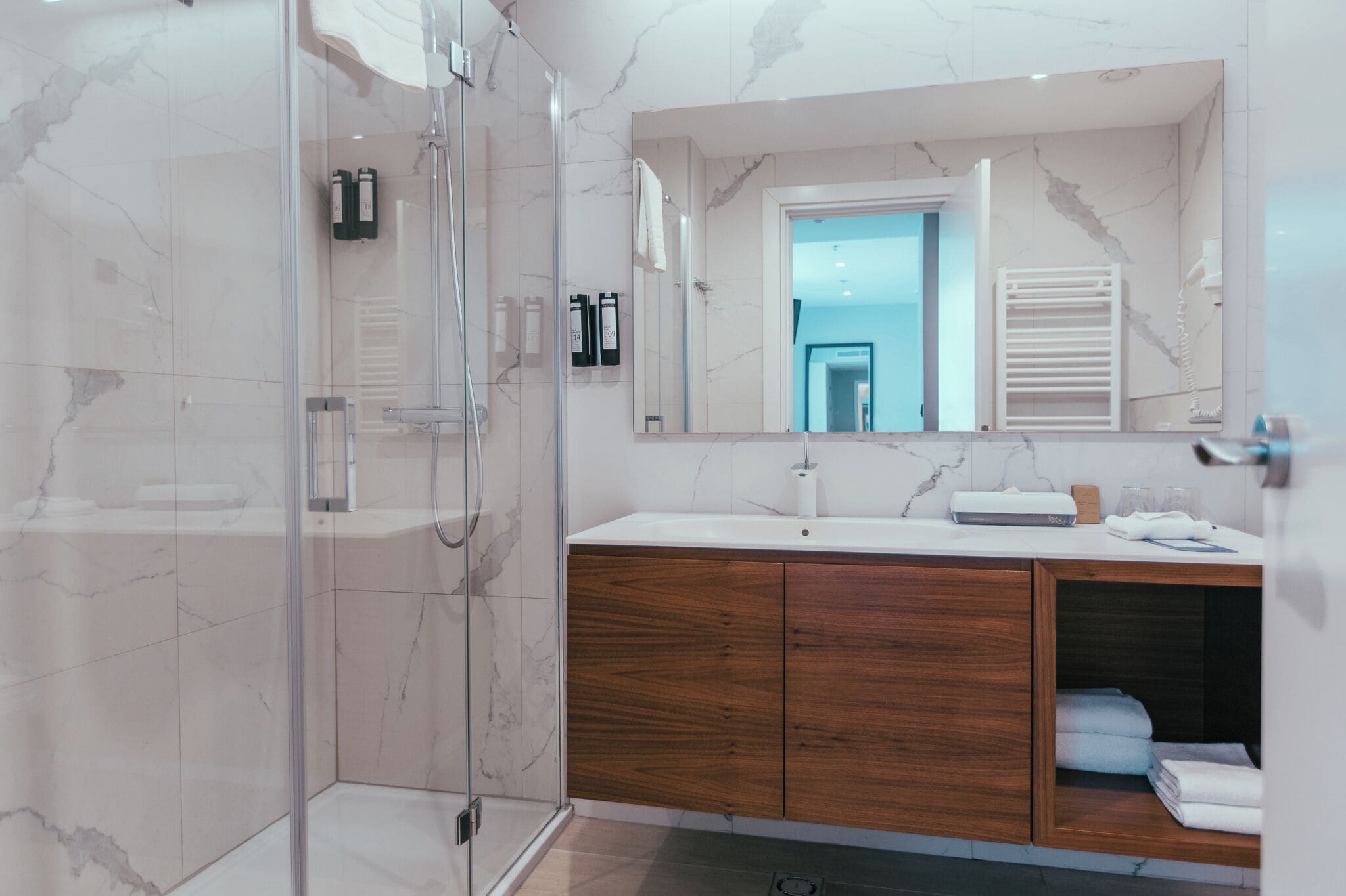 A modern bathroom with a glass-enclosed shower, marble tiles, a wooden vanity, and a large mirror.