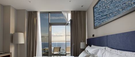 A hotel room with a large bed, a view of the sea, and a balcony with a table and chairs.