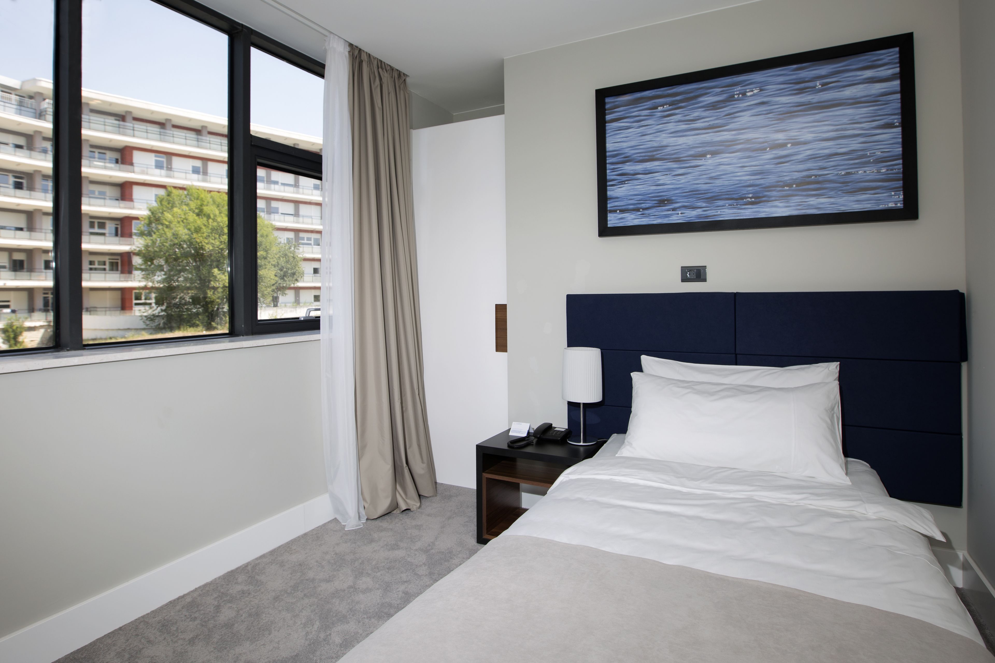 Quarto de hotel com cama, mesa de cabeceira, luminária, pintura e janela com cortinas.