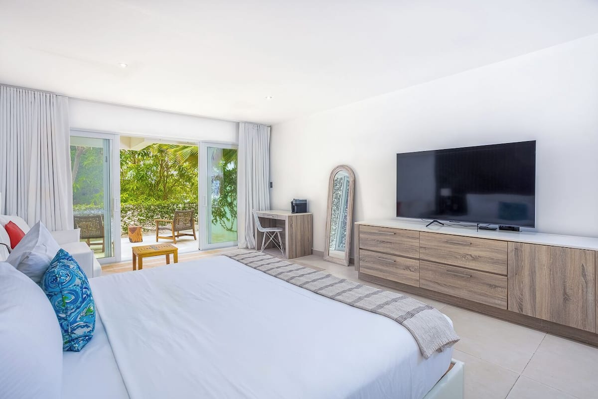 Quarto moderno com cama grande, televisão, escrivaninha e vista para o verde através da porta de vidro deslizante.