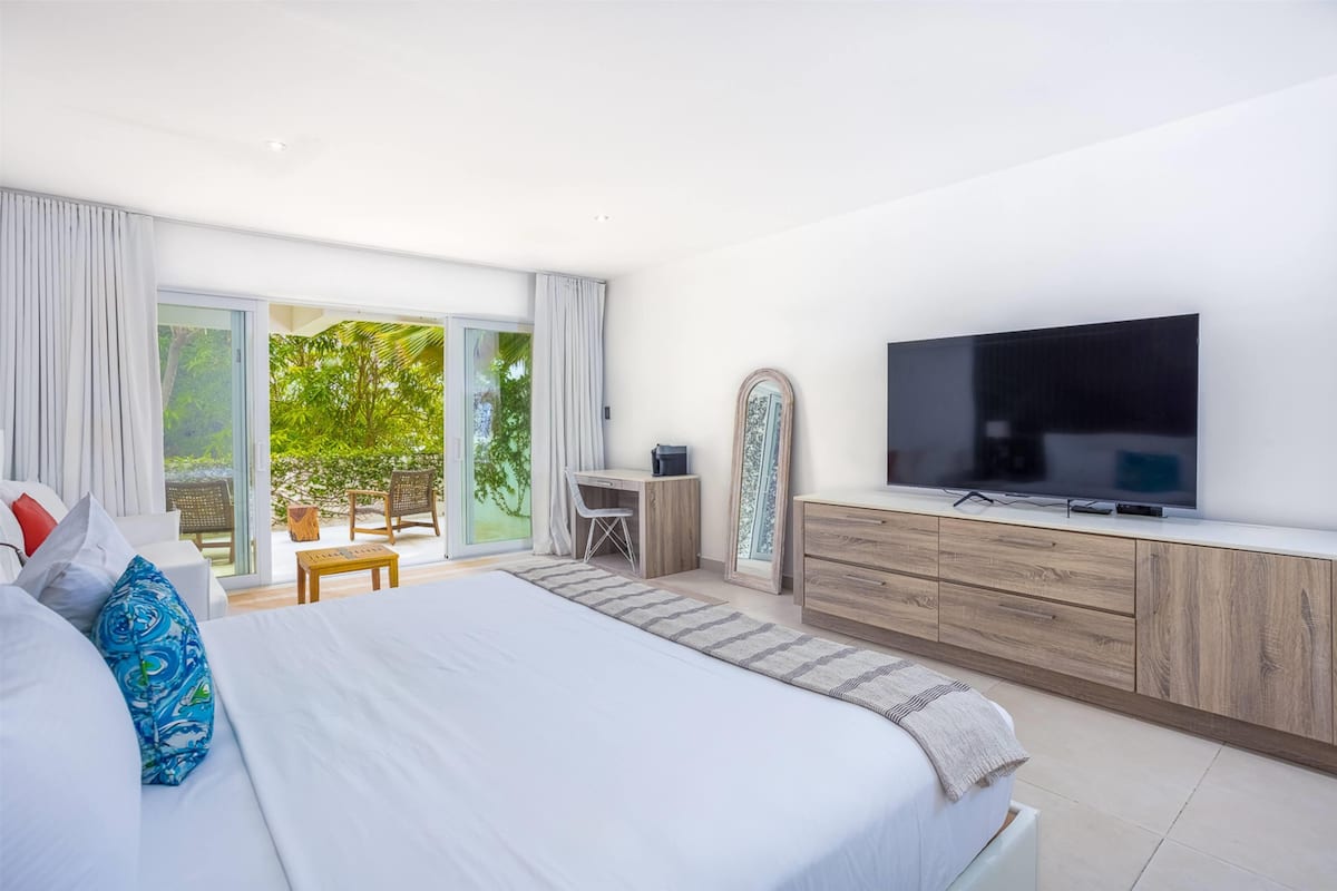 Quarto moderno com uma cama grande, televisão, escrivaninha e vista para o verde através de uma porta de vidro deslizante.