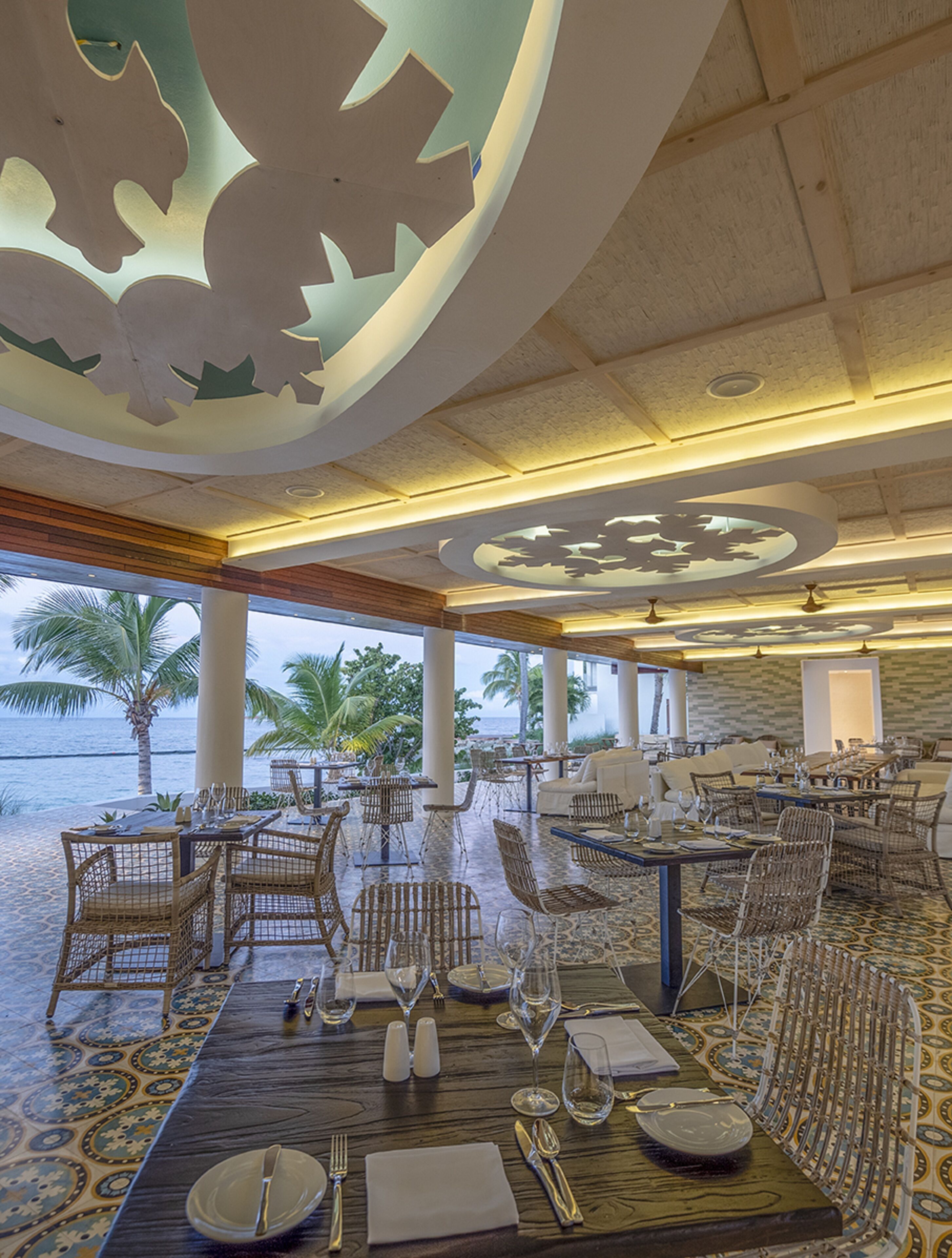 A restaurant with a view of the ocean, featuring a high ceiling with decorative cutouts, wicker furniture, and a tiled floor.
