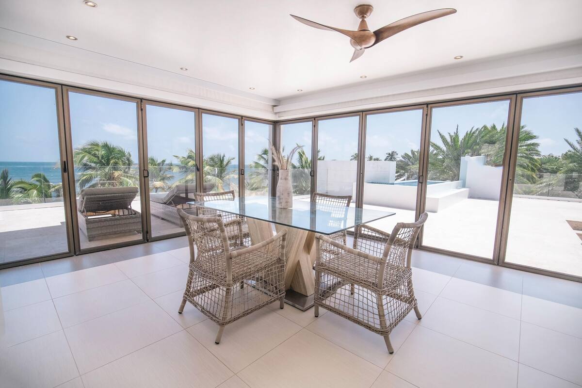 A spacious room with a glass table and wicker chairs, large windows with a view of the ocean and palm trees, and a ceiling fan.