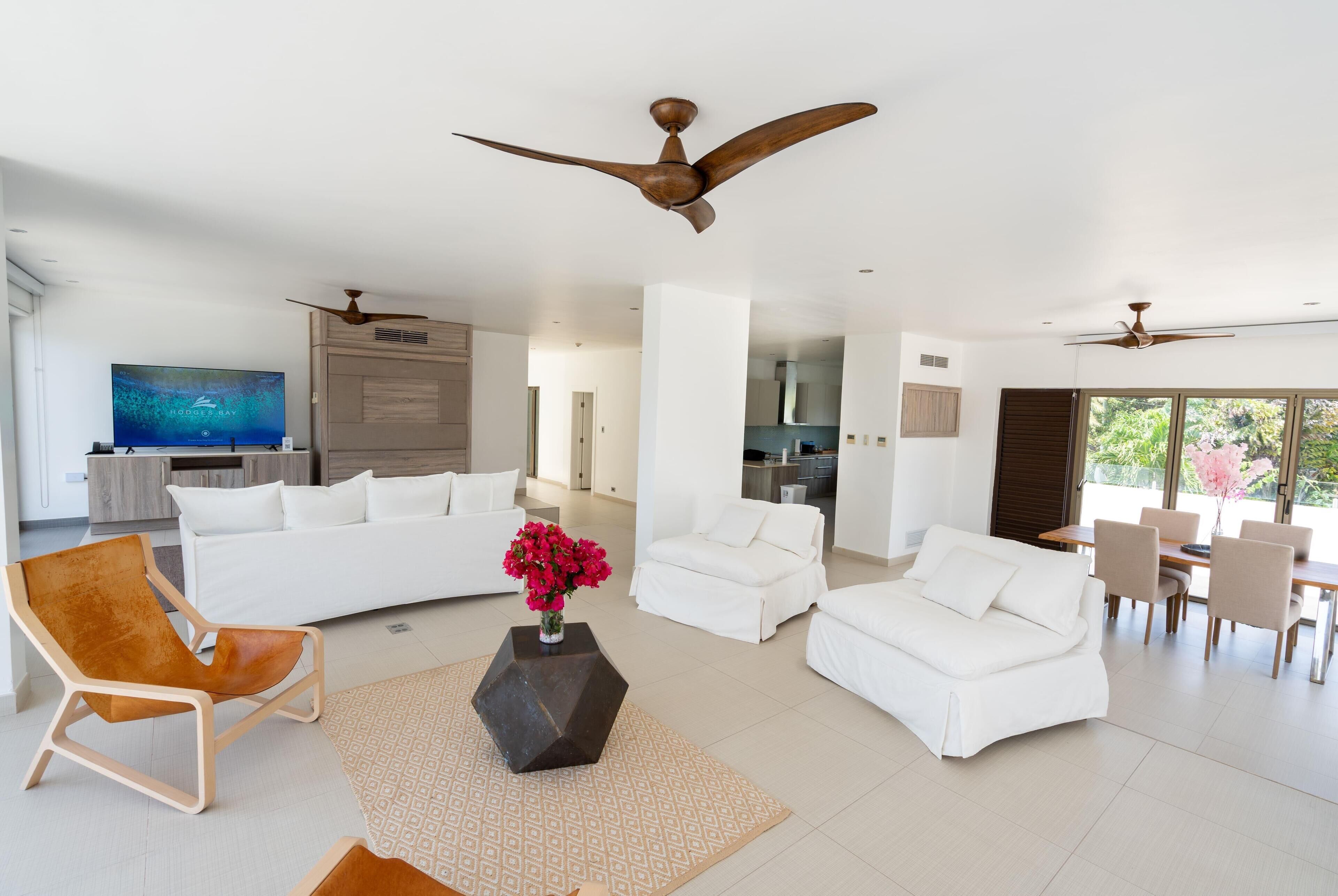 Amplia sala de estar con un televisor de pantalla plana grande, sofás blancos, una silla de madera y una mesa de comedor con sillas.