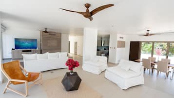 Amplia sala de estar con un televisor de pantalla plana grande, sofás blancos, una silla de madera y una mesa de comedor con sillas.
