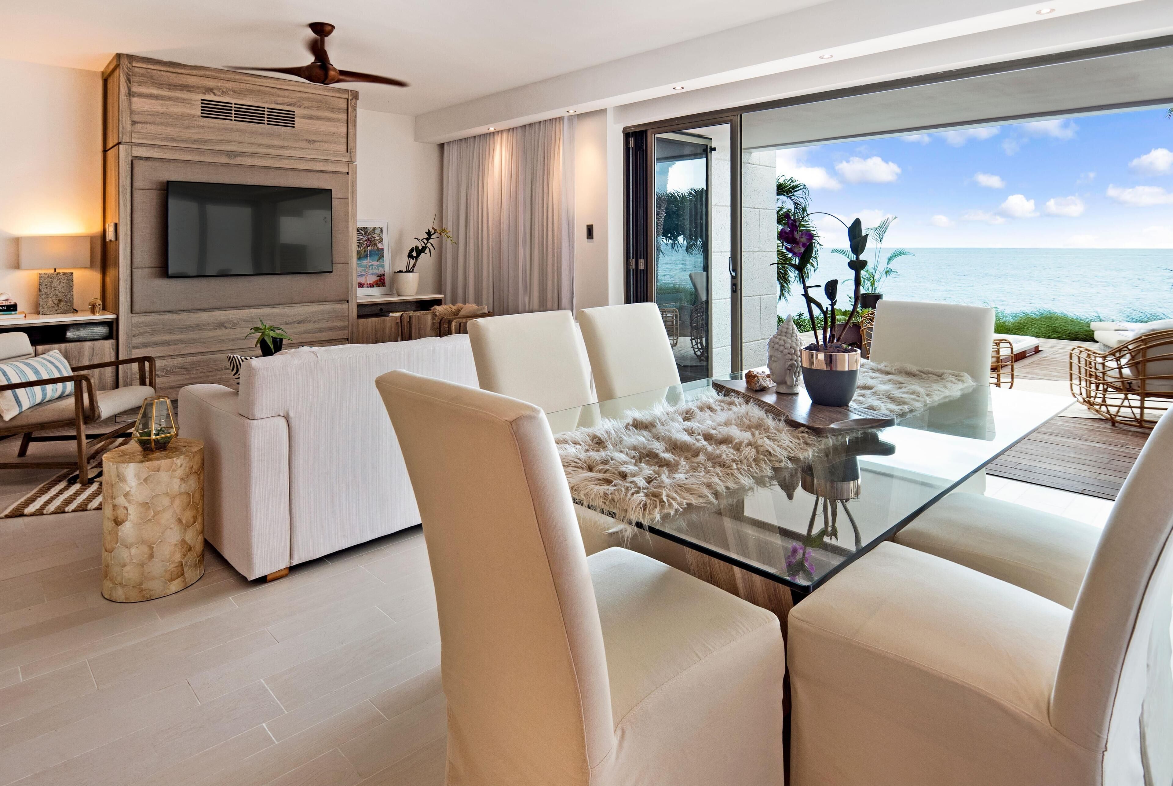 A modern living room with a glass table, white sofas, and a large TV mounted on a wooden wall.