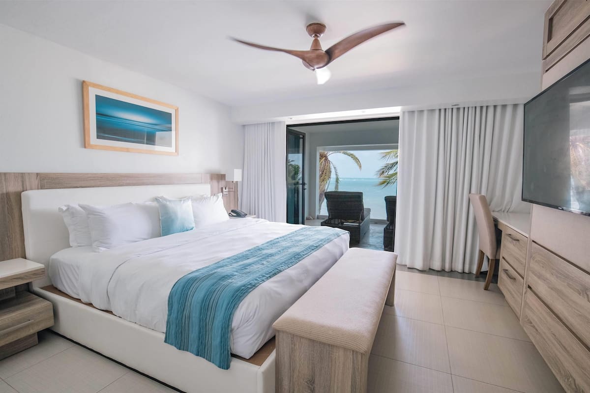 A modern hotel room with a large bed, a desk, a television, and a balcony with a view of the ocean.