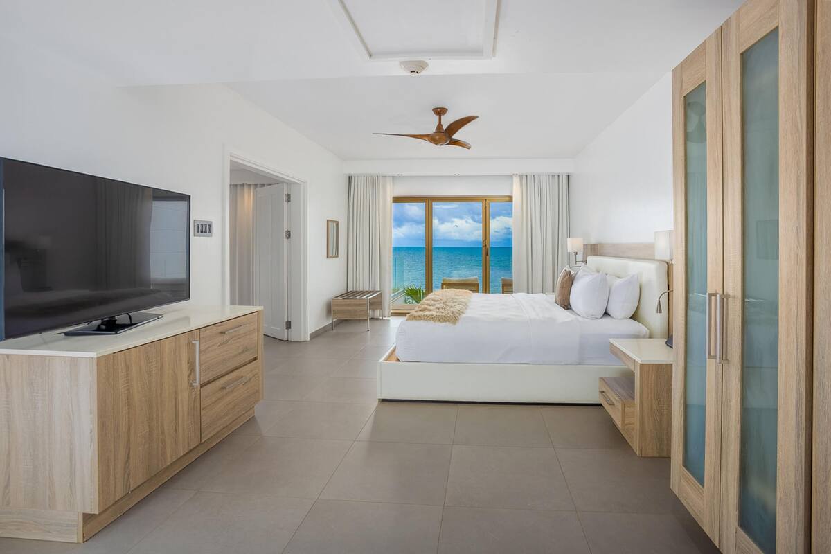 Quarto de hotel moderno com uma televisão de tela plana grande, cama com roupa de cama branca, ventilador de teto e vista para o oceano através de amplas janelas.
