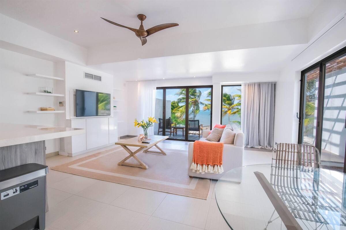 A modern living room with a sofa, coffee table, and a TV on a white wall. There is a balcony with a view of palm trees and a glass door.